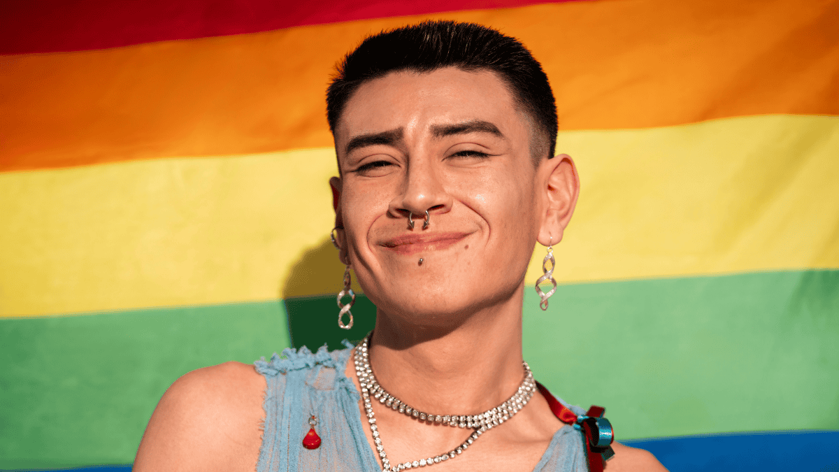 A gay man smiles in front of a rainbow flag. He's wearing a light blue top, multiple necklaces, and large hoop earrings. He has piercings on his nose and lip.