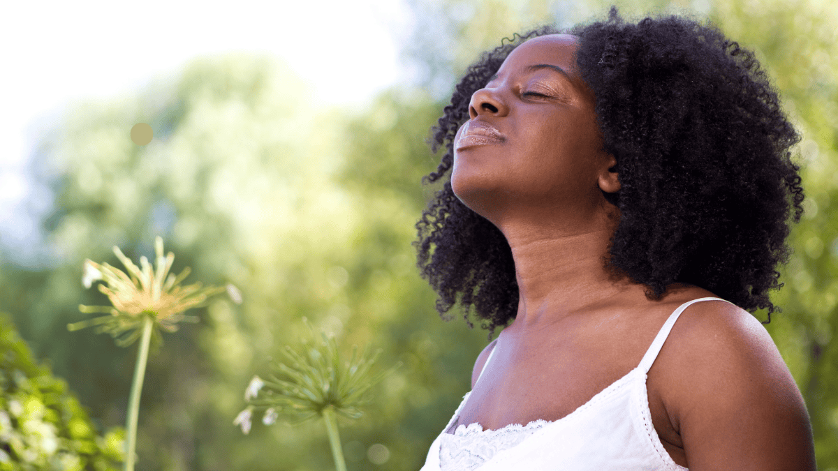 A queer woman with eyes closed and head tilted back, breathing in fresh air outdoors with blurred green foliage and flowers in the foreground.