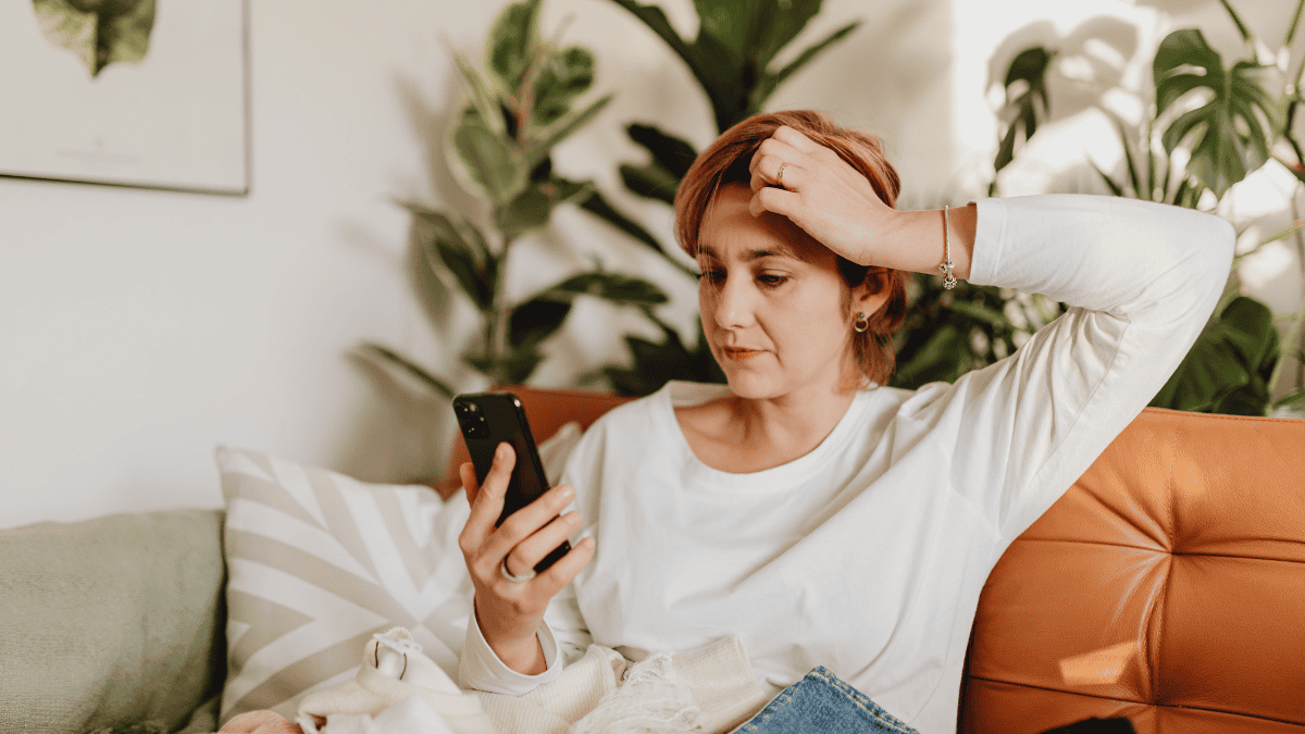 Woman looking frustrated while checking her phone, sitting on a couch at home